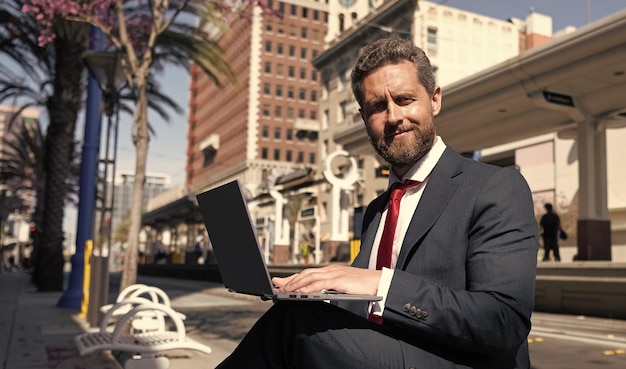 Homem de negócios maduro de terno sentado no banco na cidade ao ar livre trabalhando online em redes de laptop