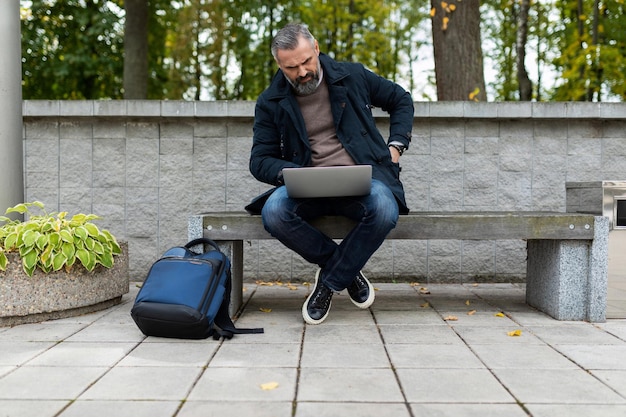 Homem de negócios maduro trabalhando online em um laptop sentado em um banco em um parque da cidade