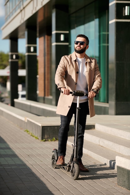 Homem de negócios moderno e elegante em óculos de sol, casaco e calças, movendo-se em uma scooter elétrica ao longo da parte externa de um grande centro de escritórios contemporâneo
