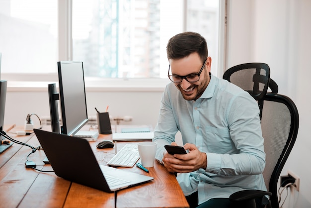 Homem de negócios novo alegre na mensagem da leitura do escritório no telefone.