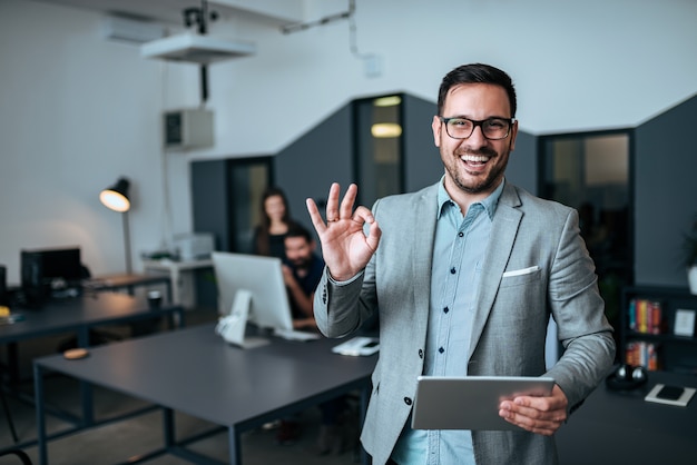 Homem de negócios novo considerável que mostra o gesto APROVADO no escritório moderno.