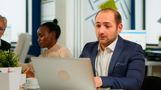 Homem de negócios ocupado usando laptop digitando sentado à mesa de conferência em sala ampla, concentrado no trabalho, enquanto diversos colegas trabalhando em segundo plano. Tecnologia moderna, conceito de ocupação de jovens.