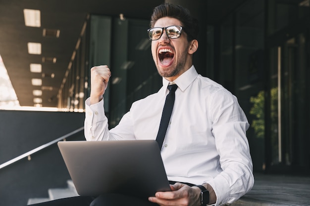 Homem de negócios perto do centro de negócios, usando o computador portátil, fazer o gesto de vencedor.