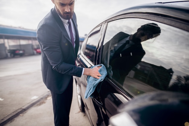 Homem de negócios que lustra seu carro com pano de microfibra na frente da lavagem de carros.