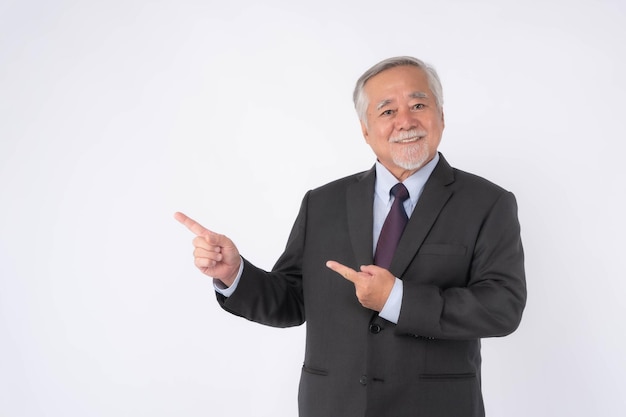 Homem de negócios sênior asiático com terno sente-se feliz, boa saúde, apontando o dedo para o espaço vazio da cópia para o produto presente ou espaço em branco para publicidade isolada no fundo branco