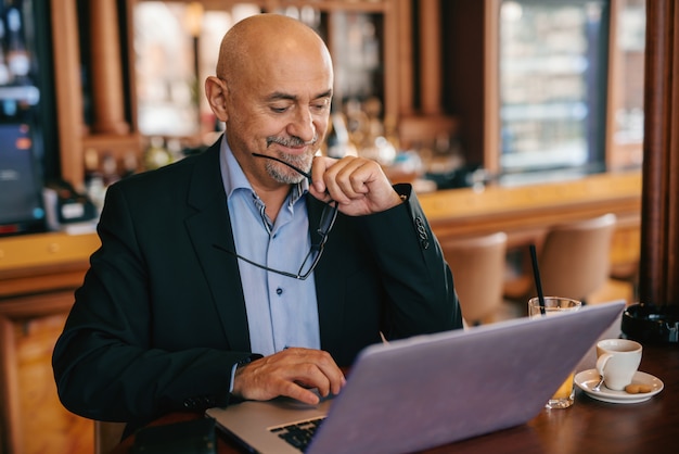 Homem de negócios sênior barbudo de terno segurando óculos na mão e sorrindo enquanto estiver usando o laptop na cafeteria.