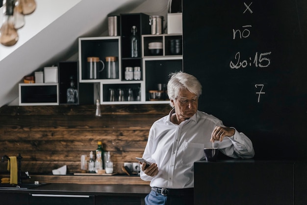 Homem de negócios sênior com smartphone e xícara de café na cozinha