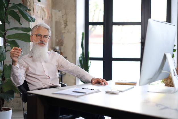 Homem de negócios sênior com uma barba estilosa, trabalhando no computador em sua mesa de escritório.