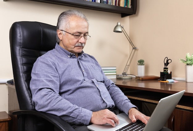 Homem de negócios sênior trabalhar em casa. Um homem idoso de óculos está trabalhando remotamente usando um laptop. Trabalho remoto durante o conceito de coronovírus