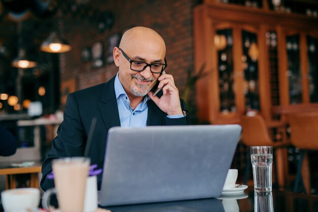 Homem de negócios sênior usando telefone inteligente e laptop enquanto está sentado no café