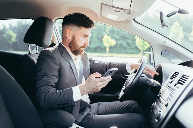 Homem de negócios sentado ao volante de um carro, frustrado, não consegue entender a direção e as instruções do carro funky, lê informações adicionais no telefone