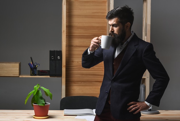 Homem de negócios sério bebendo café no escritório. Café quente. Drinks. Retrato do empresário barbudo confiante. Homem de negócios sério de fato. Trabalhador de escritório barbudo bonito.