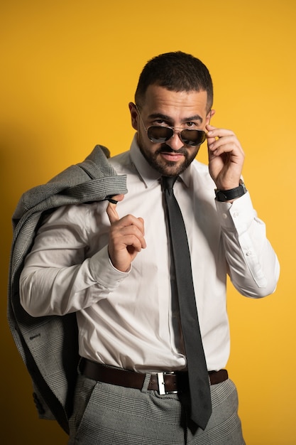 Homem de negócios sério e barbudo vestido em suíte acinzentada posando segurando sua jaqueta no ombro pendurando-a atrás, olhando baixando os óculos de sol isolados no fundo amarelo.