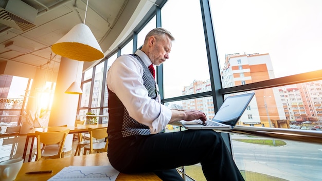 Homem de negócios sério sênior sentado perto da janela com vista panorâmica da cidade laptop nas mãos homem pensando enquanto verifica e-mails