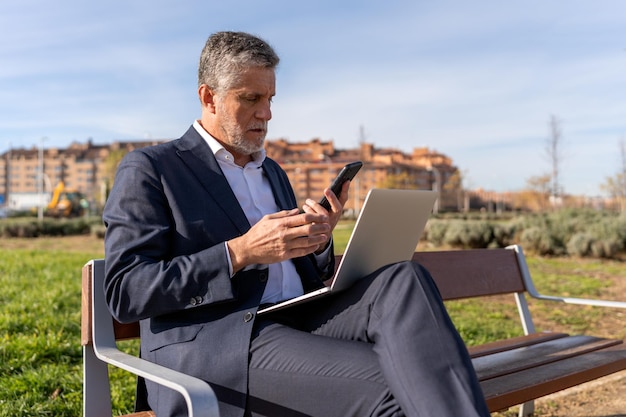 Homem de negócios sério verificando smartphone durante o trabalho no laptop