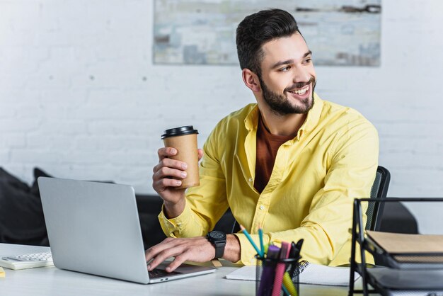 Foto homem de negócios sorridente segurando um copo de papel usando um laptop e olhando para longe