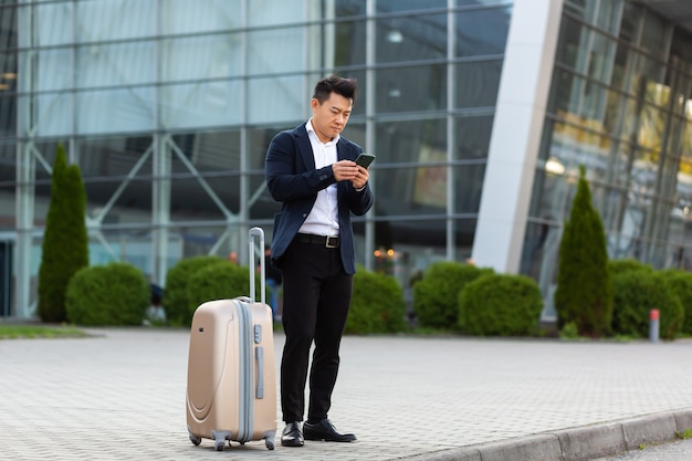 Homem de negócios tentando chamar um táxi usando um aplicativo e um telefone celular, um homem asiático na estação de trem com uma mala grande