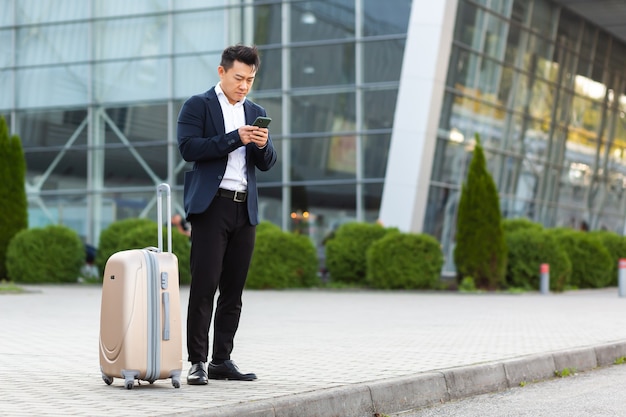 Homem de negócios tentando chamar um táxi usando um aplicativo e um telefone celular, um homem asiático na estação de trem com uma mala grande