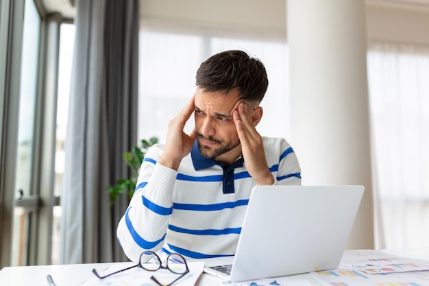 Homem de negócios trabalhando em escritório com dor de cabeça dolorida da mesa trabalhando e sentado o dia todo usando computador portátil ou notebook sofrendo dor de cabeça trabalhador doente conceito de excesso de trabalho