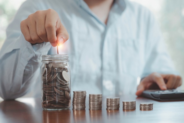 Homem de negócios usando calculadora e colocando moeda para salvar jar e empilhar moedas, economia de dinheiro do depósito e conceito de crescimento do lucro do negócio.