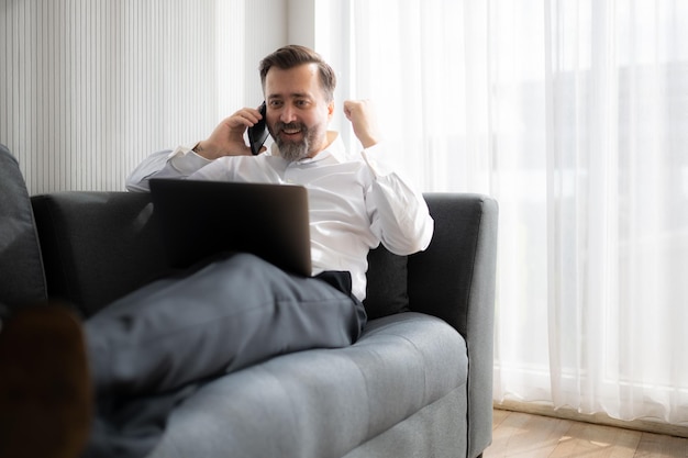 Homem de negócios usando laptop e falando no telefone enquanto está sentado no sofá em casa