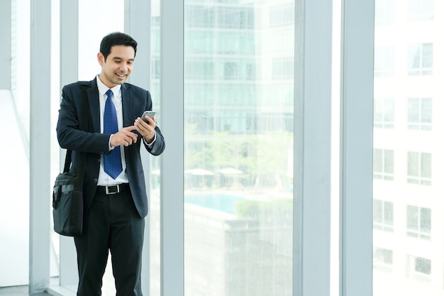 Homem de negócios usando o telefone inteligente com um rosto sorridente feliz ao caminhar pela janela