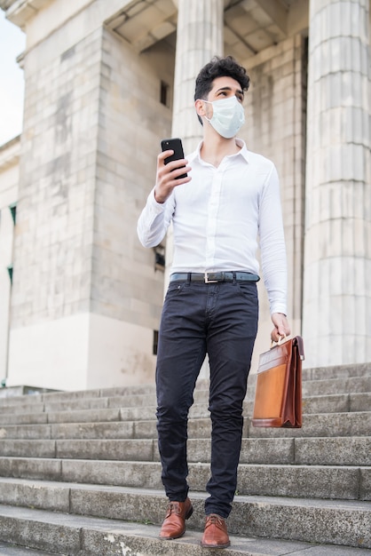 Homem de negócios usando uma máscara facial e usando seu telefone celular enquanto caminha ao ar livre