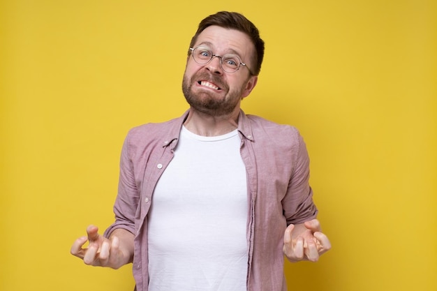 Homem de óculos redondos e camisa está estressado descobre os dentes furiosamente e aperta os dedos com força