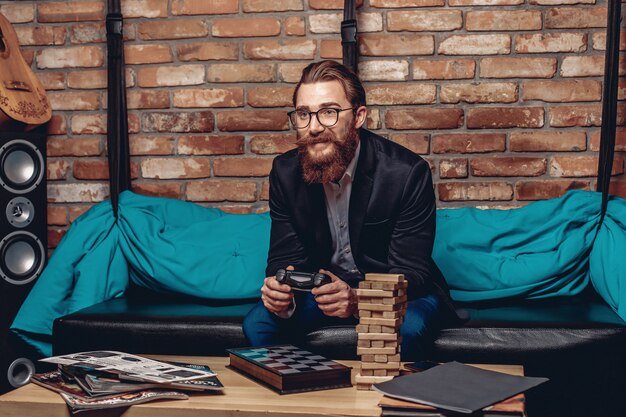 homem de óculos, sentado em um sofá e jogando um console. Jogos de tabuleiro na mesa.