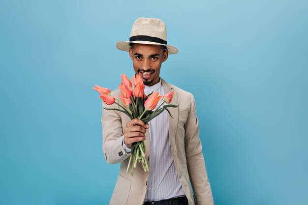 Homem de olhos castanhos com chapéu segurando tulipas cor de rosa na parede azul