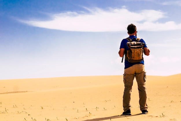 Homem de pé atrás com uma mochila olhando as dunas do deserto de areia e o céu azul