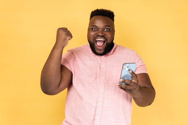 Homem de pé com celular nas mãos e punho cerrado comemorando sua vitória olha para a câmera