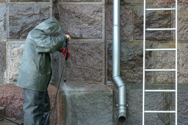 Homem de pé em uma escada lava as paredes do prédio com um jato de água sob pressão Traje de proteção química Descontaminação do prédio do coronavírus Medidas de segurança do covid19