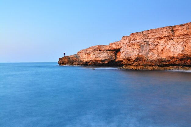 Homem de pé na rocha ao longo da costa do mar negro, com águas azuis claras e a paisagem de céu azul na crimeia.