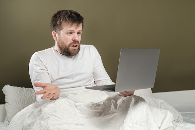 Homem de pijama trabalha com laptop remotamente sentado na cama e fala emocionalmente por videochamada