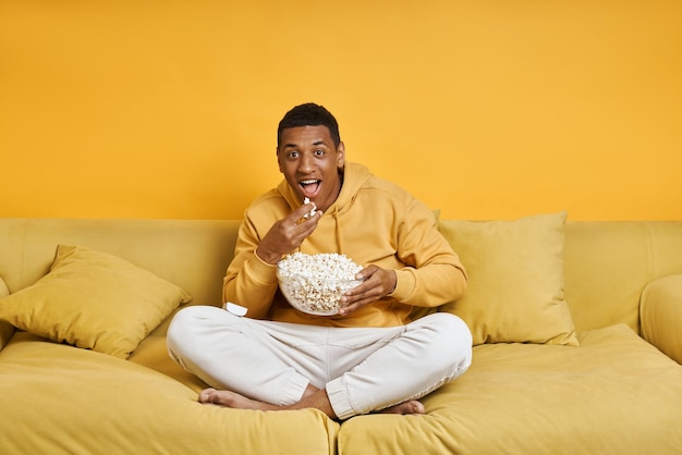 Homem de raça mista feliz com lanches enquanto relaxa no sofá com fundo amarelo