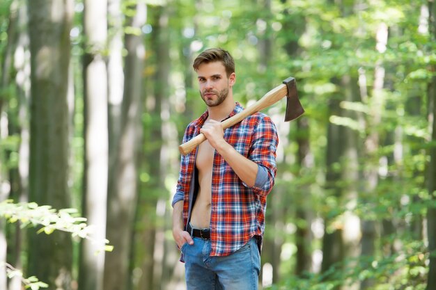 Homem de rancho madeireiro moderno carrega machado cara sexy veste camisa desabotoada na floresta poder masculino e lenha de energia no acampamento acampar e caminhar atividade ao ar livre homem forte com machado lenhador