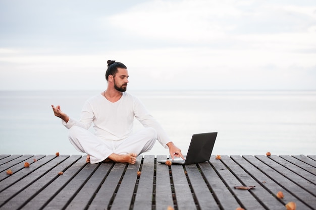 Homem de roupas brancas meditando ioga com laptop no píer de madeira