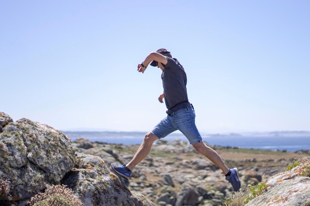 Foto homem de roupas de verão a saltar entre as rochas em frente ao mar