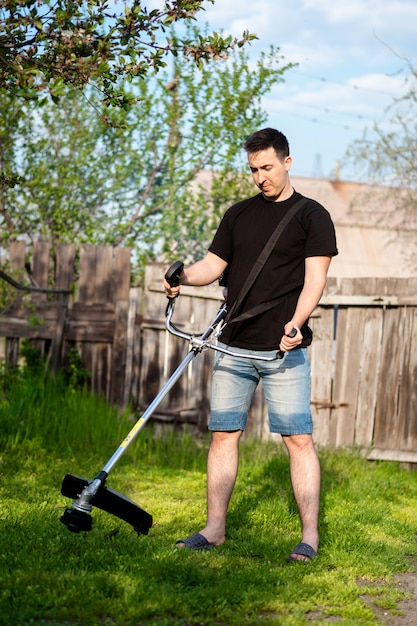 Homem de short com um cortador de escova profissional, cortar a grama no quintal. gramado verde, cerca velha como pano de fundo. tempo ensolarado de primavera.