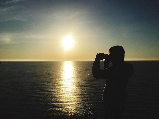 Foto homem de silhueta fotografando o mar contra o céu durante o pôr do sol