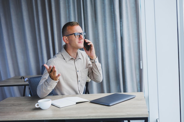 Homem de sucesso falando no smartphone e navegando no netbook enquanto navega na internet no escritório à mesa