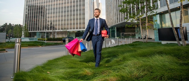 Homem de sucesso feliz em trajes formais com sacolas de compras e caixa de presente fora do escritório, negócios.