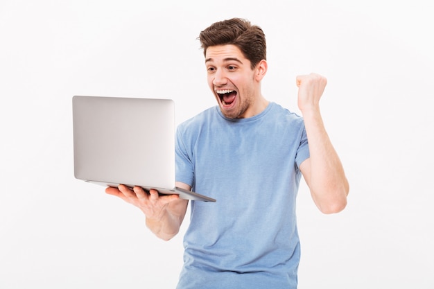 Homem de sucesso feliz no punho de aperto de camiseta como sortudo ou vencedor enquanto trabalhava na internet no laptop, isolado sobre a parede branca