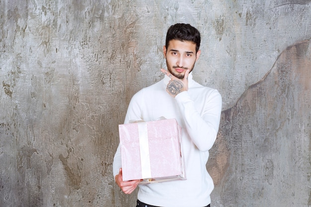 Homem de suéter branco segurando uma caixa de presente roxa embrulhada com fita e parece confuso ou pensativo.