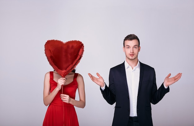 Foto homem de terno e mulher de vestido vermelho segurando um balão em forma de coração vermelho sobre o rosto em fundo branco