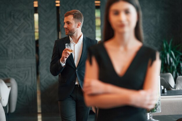 Foto homem de terno preto e com bebida nas mãos olha para mulher de vestido