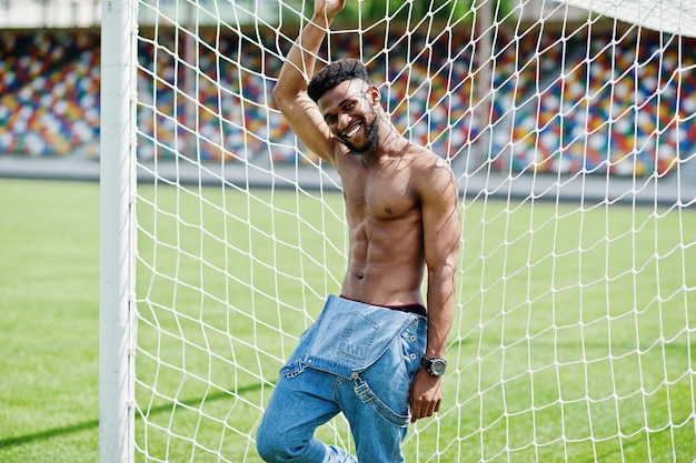 Homem de torso nu muscular afro-americano sexy bonito em macacão jeans posado na grama verde contra portões de futebol do campo do estádio Retrato de homem negro na moda