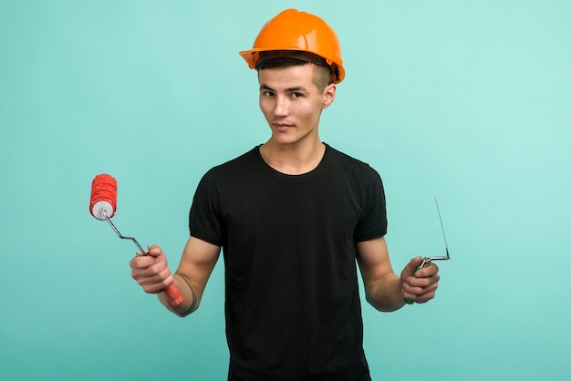 Homem de trabalho asiático em um capacete laranja está com um rolo de pintura e uma tralha em uma imagem de fundo azul