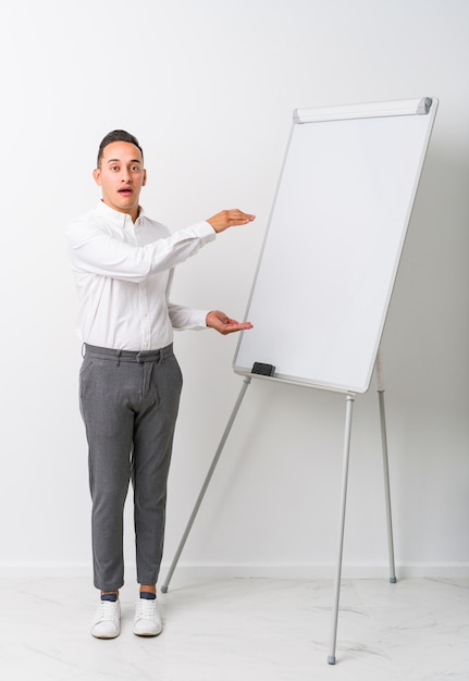 Homem de treinamento latino novo com um quadro branco isolado chocado e espantado segurando um espaço de cópia entre as mãos.